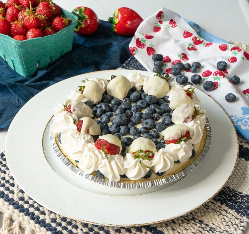 berry1 photo of a whole pie on a white platter, quart of strawberries in background, the pie has a center of fresh. blueberries surrounded by whipping cream and white chocolate covered strawberries as garnish