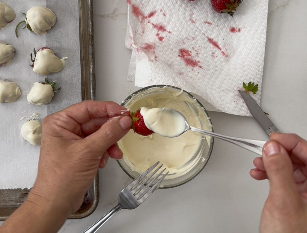 bowl of melted white chocolate with fresh strawberries person is spooning melted chocolate over the strawberry halves and placing them on a parchment covered baking sheet