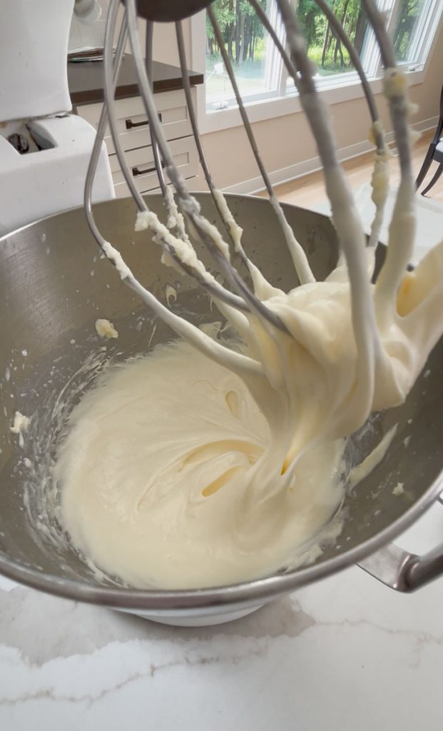stand mixer with the whisk attachment pulled up and cheesecake filling dripping from it into the bowl
