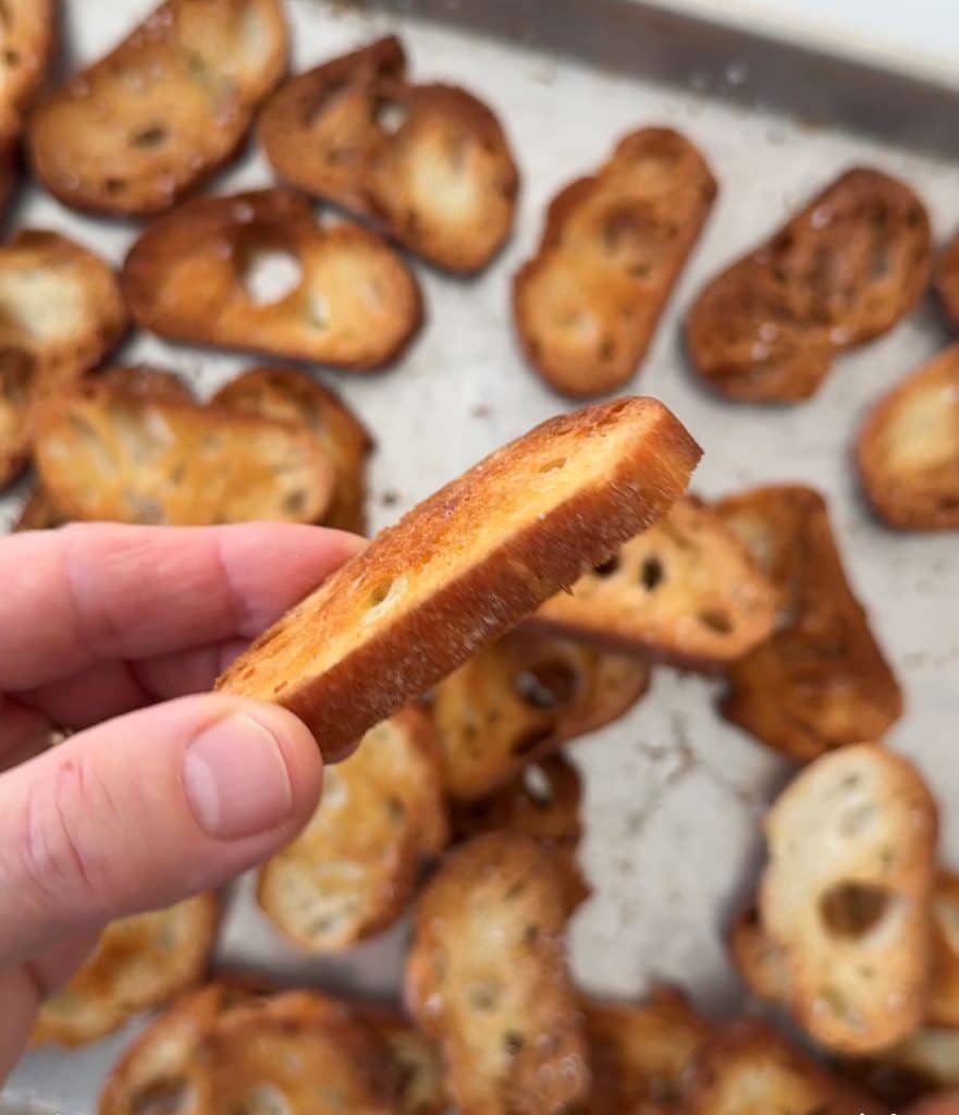 fingers holding a baked crostini over the baking sheet of crostini