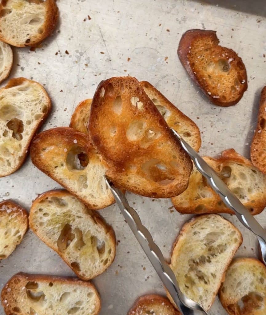 tongs holding a baked crostini over a baking sheet