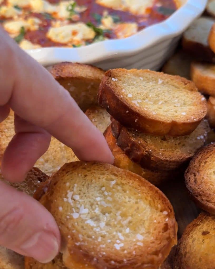 fingers picking up a crostini with a an appetizer in the background