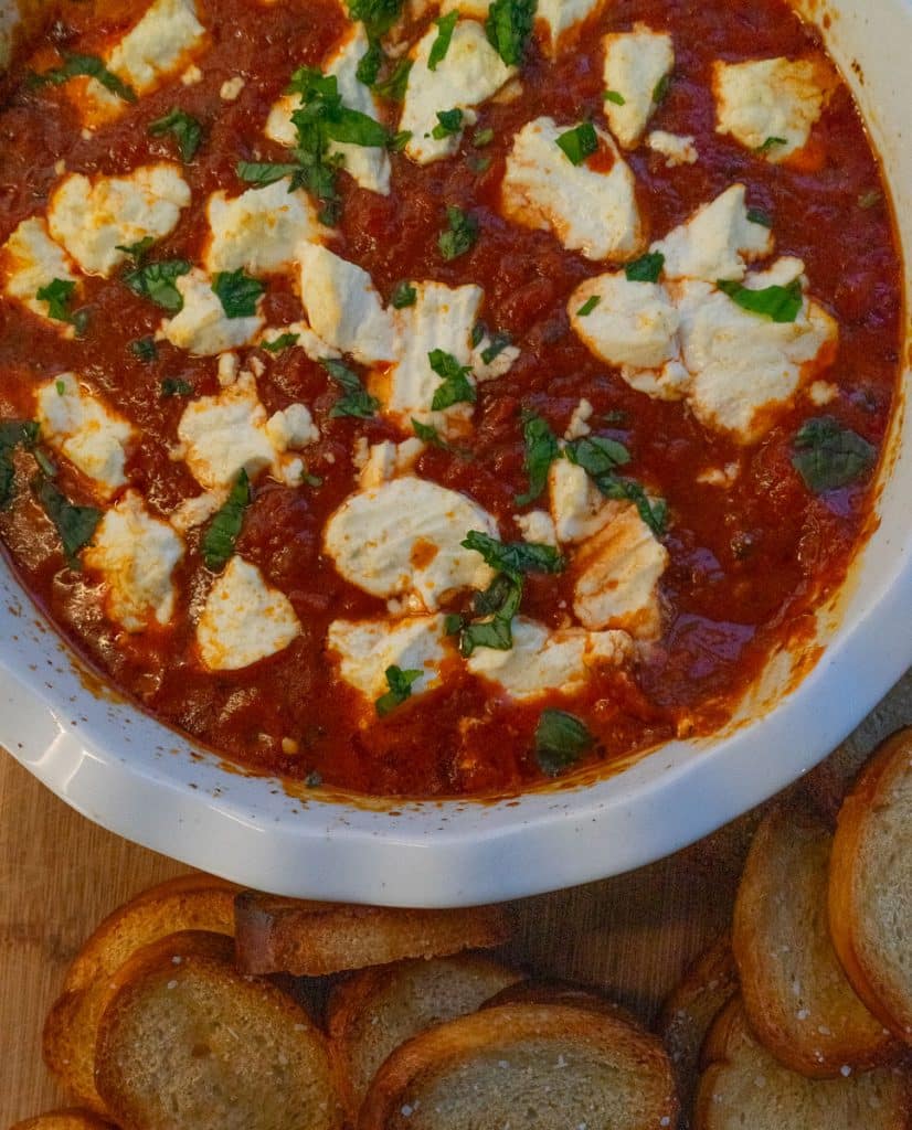white dish with marinara and goat cheese dip surrounded with crostini on a wood serving board