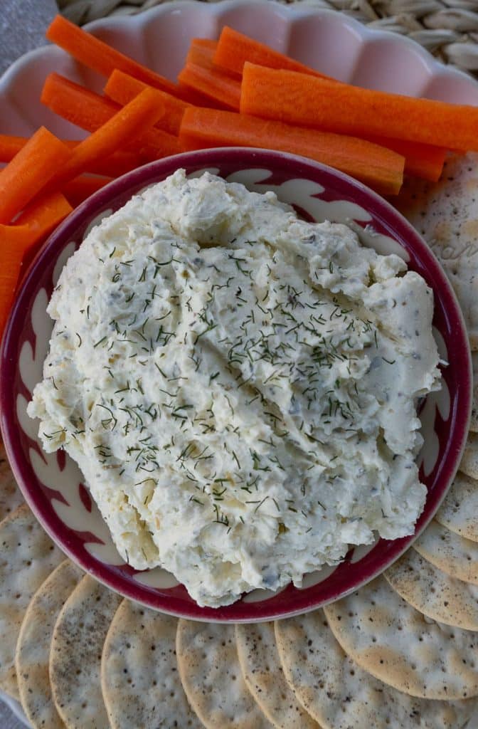close up of small red rimmed dish with feta herb dip surrounded by crackers and carrots