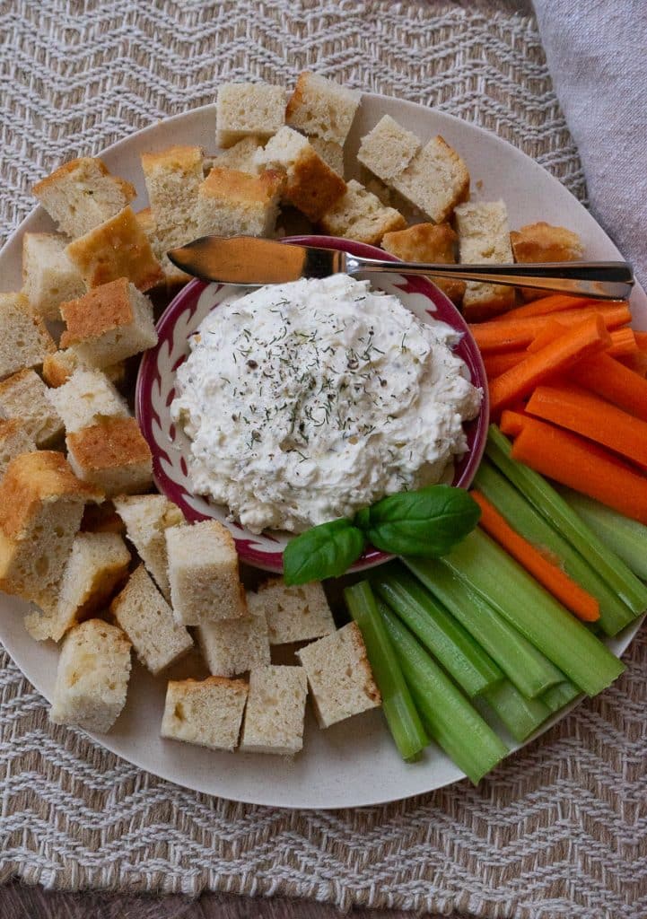 dish of feta herb dip in the middle of a plate of chunks of bread, carrots, celery on a tan placemat