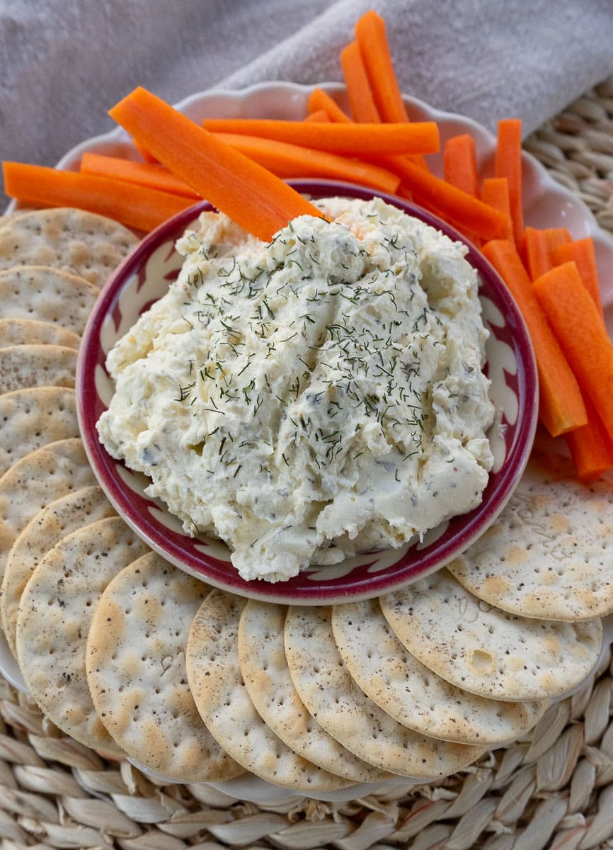 dish of feta herb dip in the middle of crackers and carrots on a plate on a rattan placemat