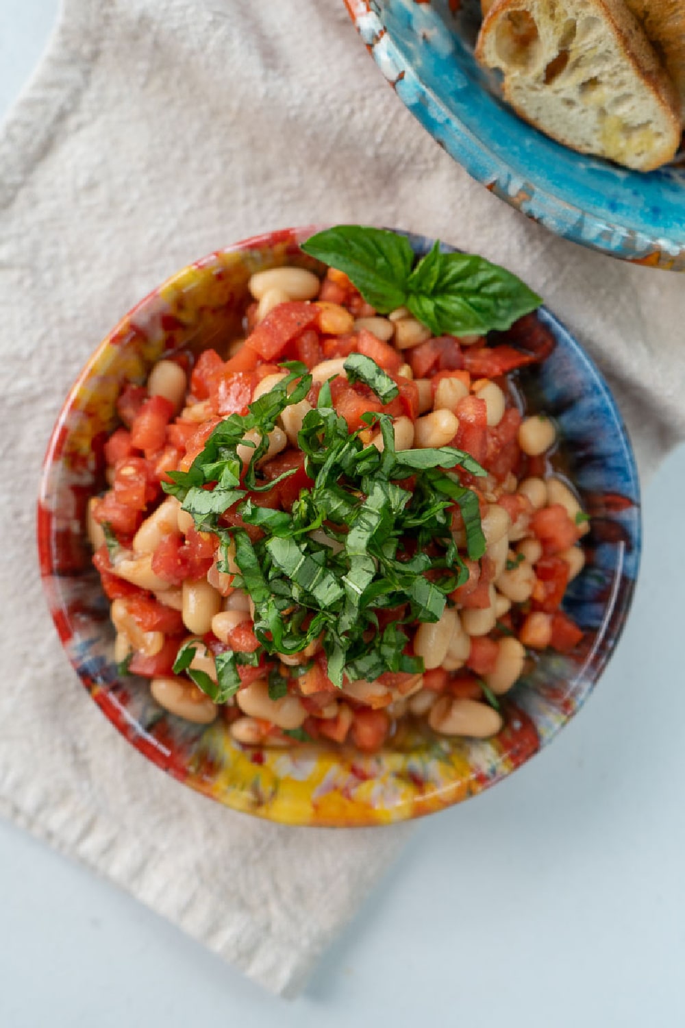 colorful ceramic bowl with bruschetta topped with fresh basil in it on a tan cloth, bowl of crostini in upper right corner