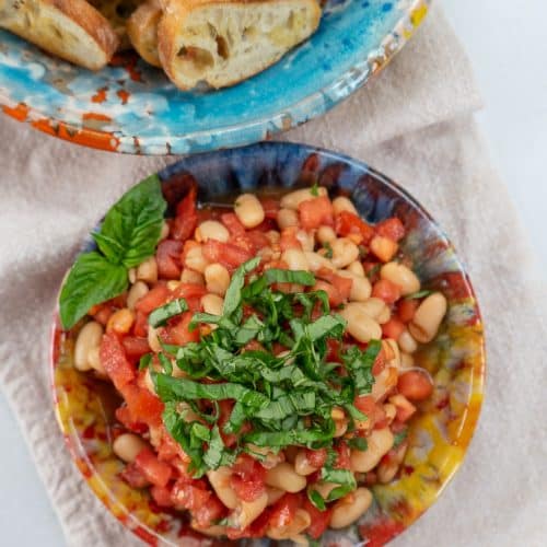 small colorful bowl full of bruschetta topped with fresh basil, next to another bowl of crostini on a tan cloth napkin