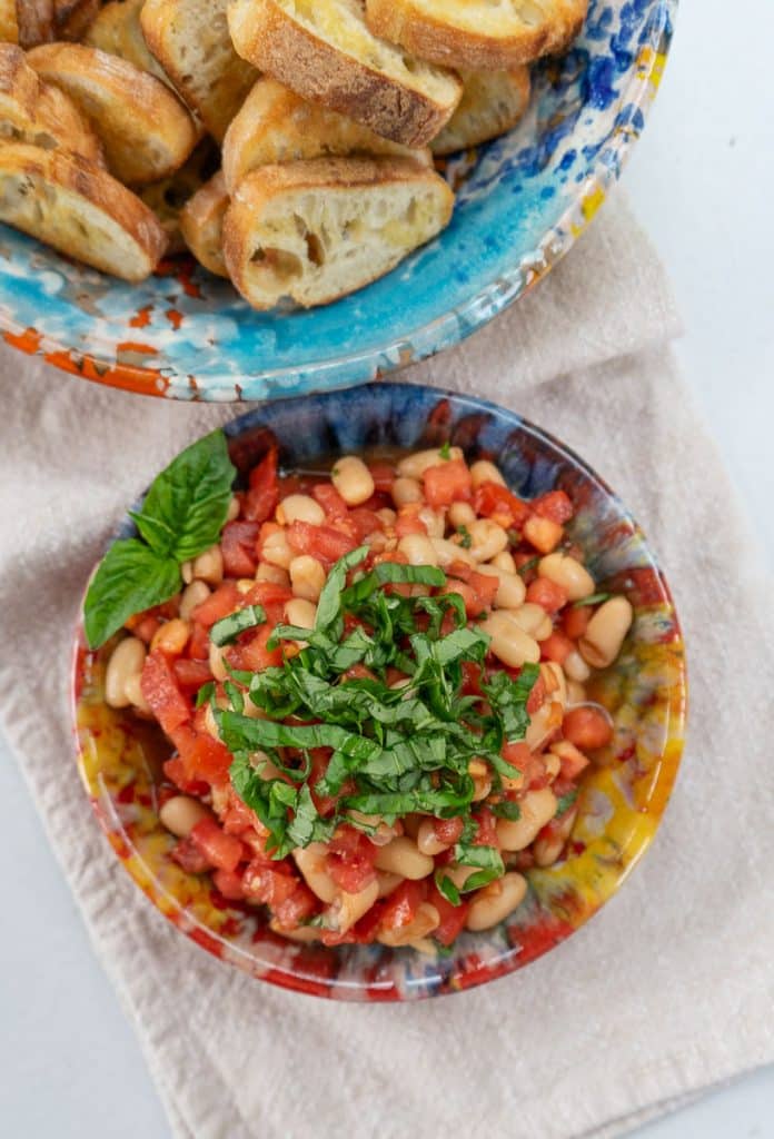 small colorful bowl full of bruschetta topped with fresh basil, next to another bowl of crostini on a tan cloth napkin