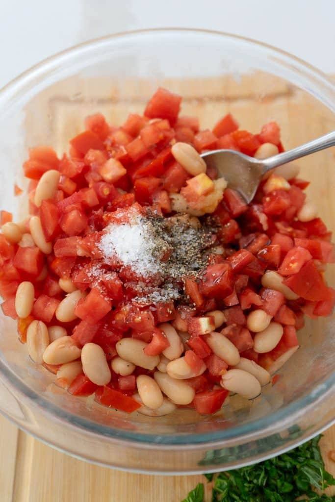 bowl with tomato, cannellini beans, olive oil, salt and pepper in it, a spoon is stirring the ingredients