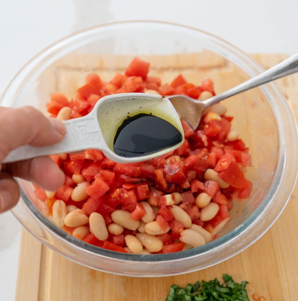 adding balsamic vinegar to the other ingredients. hand is holding a measuring spoon of vinegar over the glass bowl on a wood cutting board