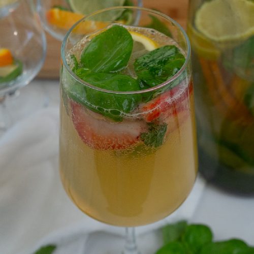 glass of basil citrus white wine sangria sitting on a white countertop with a pitcher of sangria in the background