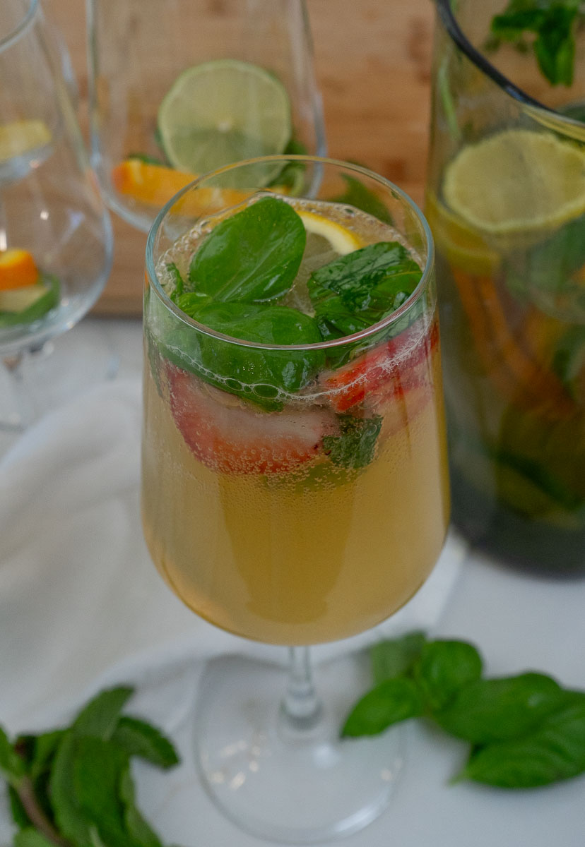 a wine glass on a white countertop with white basil citrus sangria in the glass with basil around the glass and in the glass as garnish, pitcher of sangria in the background