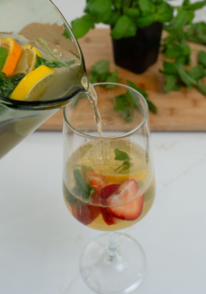 a wine glass on a white countertop with a pitcher of sangria fill the glass from the left side , cutting board with basil plant in the background