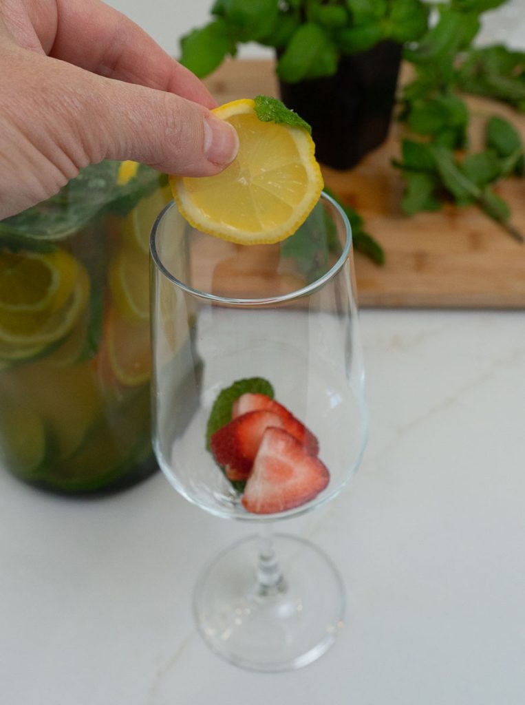 strawberries in the bottom of a long stemed wine glass a hand is dropping in a slice of lemon 