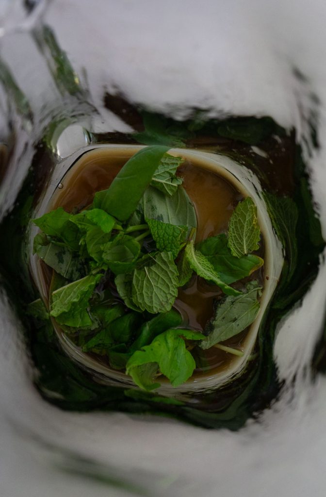 basil and mint being muddled with wine in the bottom of a glass pitcher