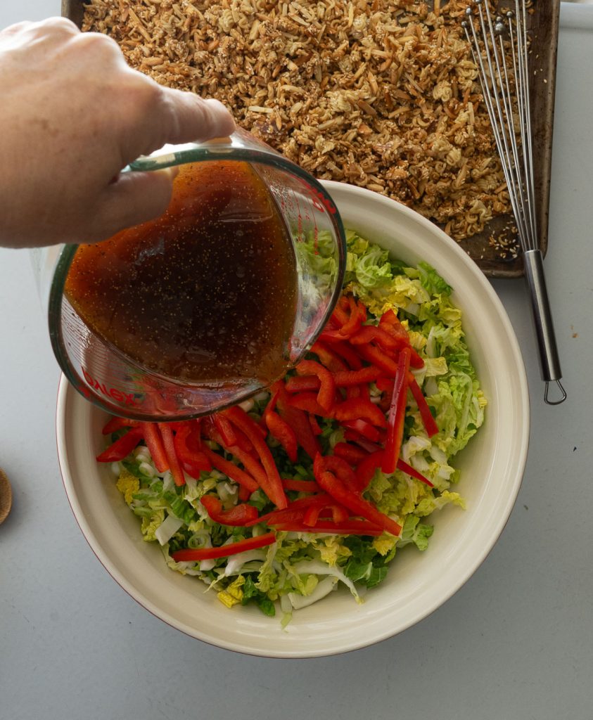 salad ingredients in bowl, pouring dressing, with crunch ramen on the top of photo on a baking sheet