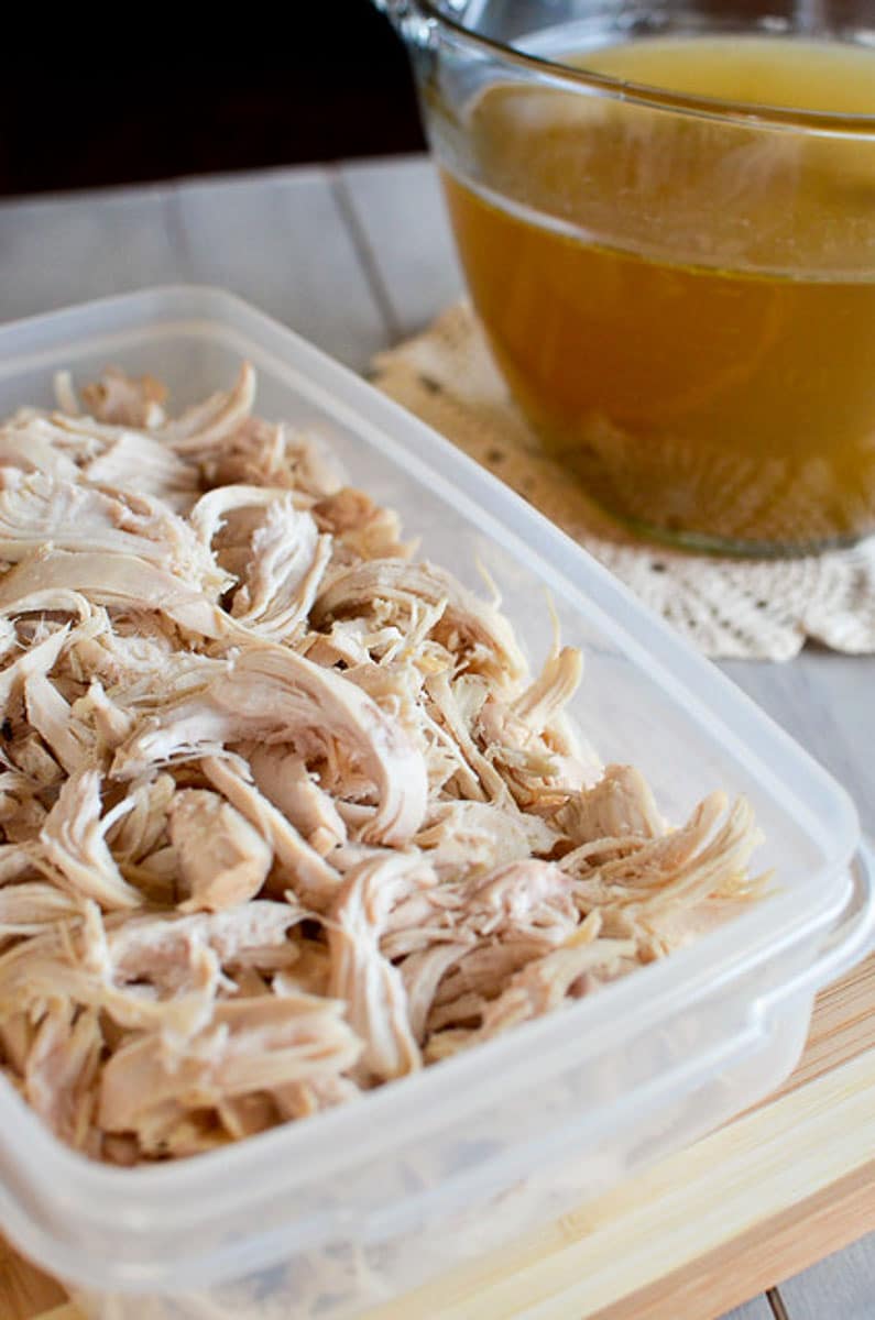 tupperware container with shredded chicken and a measuring cup of homemade broth on a table