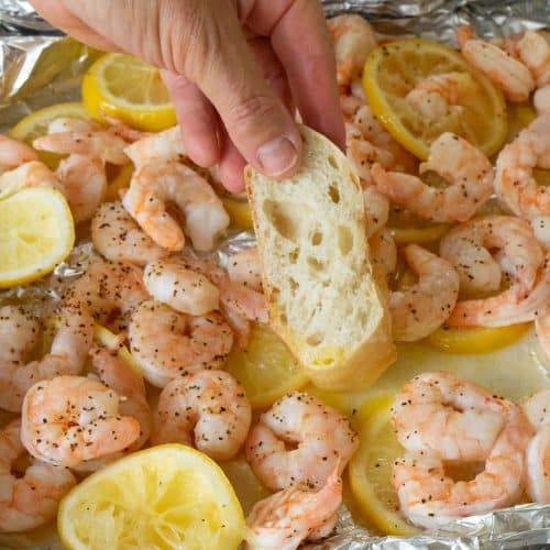 hand dipping a slice of crusty baguette into the lemon butter that is surrounding the cooked shrimp on a foil baking sheet