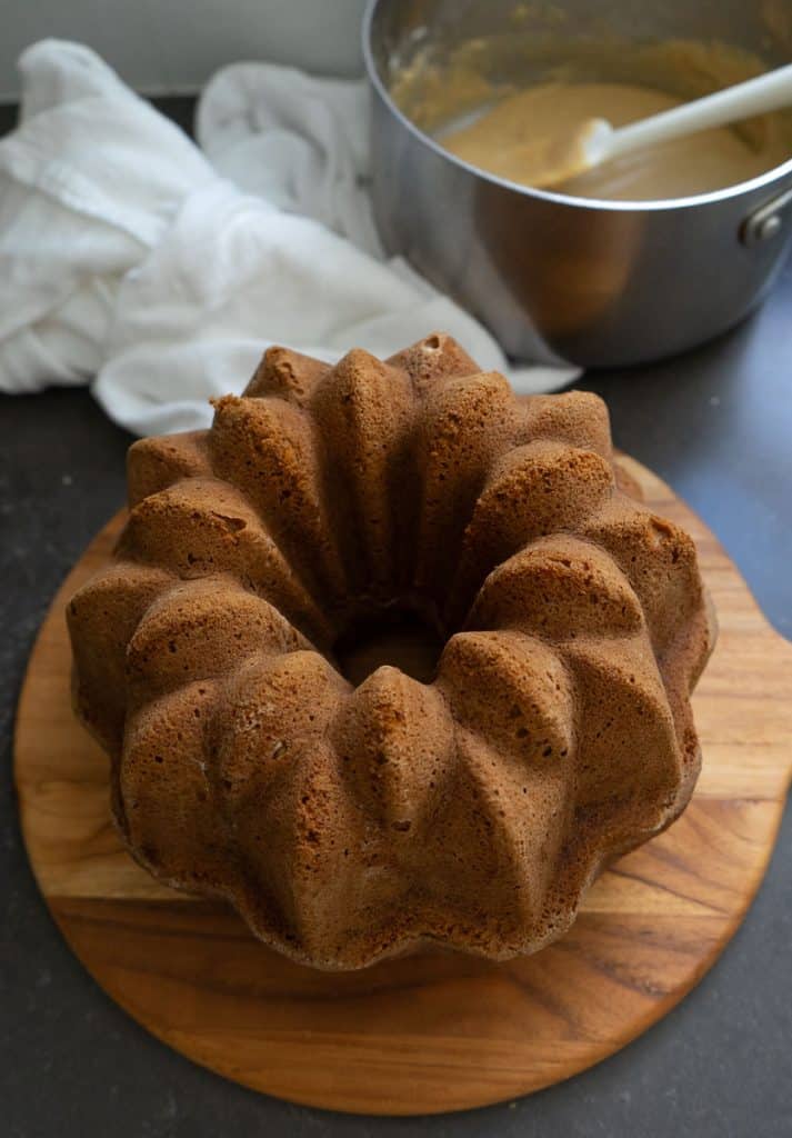 baked pumpkin bundt cake sitting on a wood tray, with sauce pan of caramel frosting 