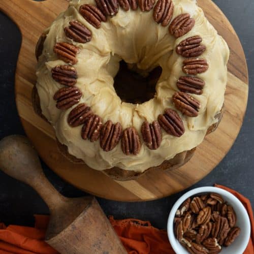 top view of pumpkin pecan bundt cake with caramel frosting on a wood platter that is sitting on a black countertop with a rolling pin and a bowl of toasted pecans next to it