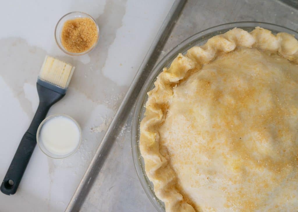 pie crust with pastry brush a small dish of milk and a small dish of turbinado sugar next to the pie. pie has been basted with milk and sprinkled with sugar