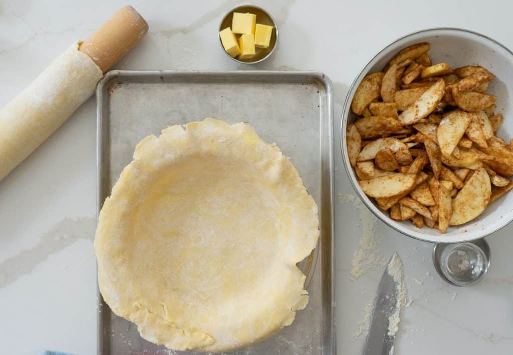 bottom crust in pie plate, apples in bowl next to pie plate, butter in a small cup above pie plate, and rolling pin with the second crust rolled out and rolled around the rolling pin
