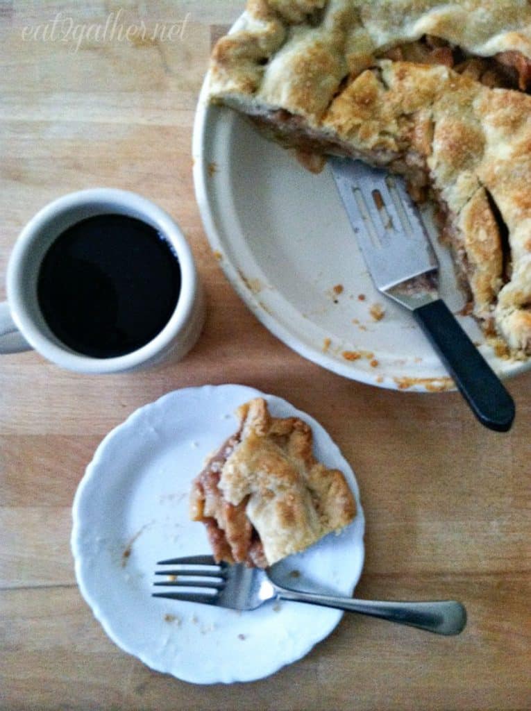 whole apple pie with a section cut out, a mug of coffee and a half eaten piece of apple pie on a white plate with a fork on the plate. there is a spatula in the pie dish all of this is sitting on a wood countertop