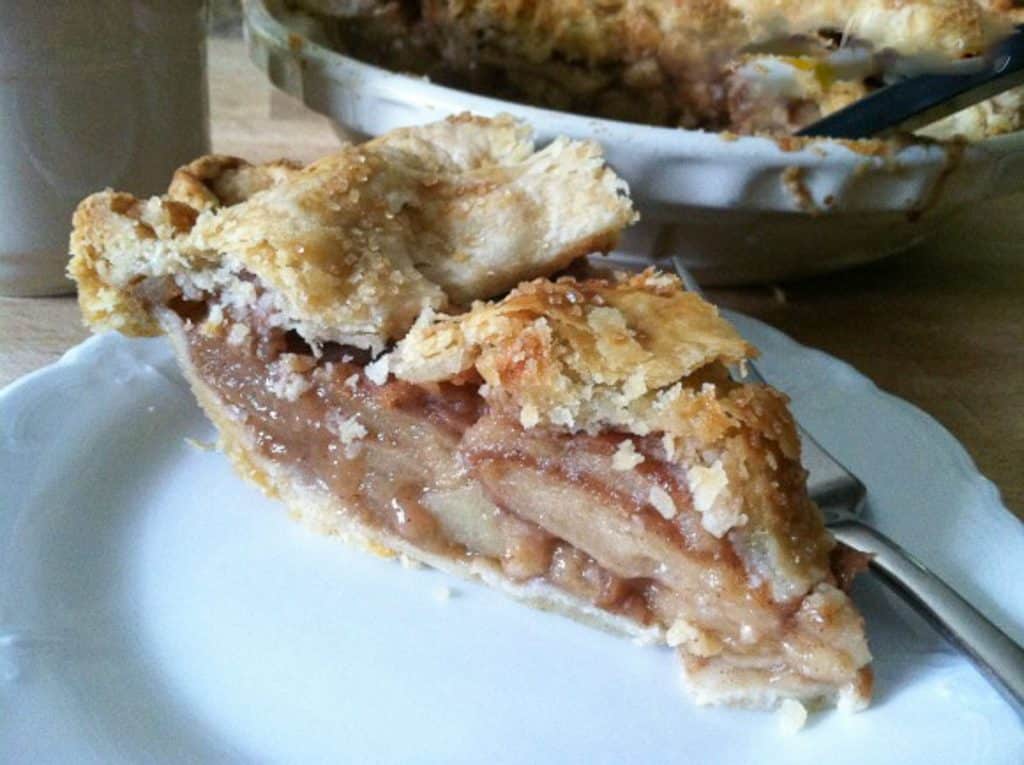 slice of apple pie on  a white plate with a fork on the plate and the pie plate with the rest of the pie in the background