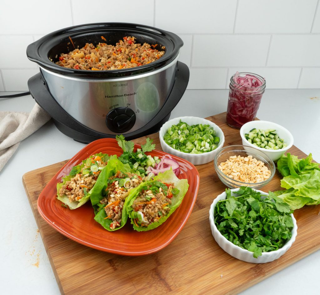 slow cooker chicken  lettuce wraps on a plate with all the toppings and crock pot in the background with meat in it