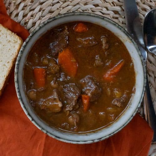 top view of stew in a bowl with piece of bread to the left side on a orange cloth