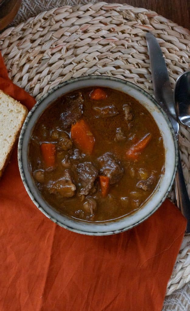 top view of stew in a bowl with piece of bread to the left side on a orange cloth