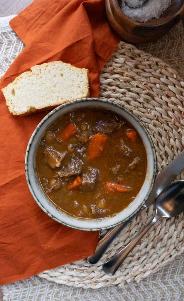 top view of stew in a bowl with piece of bread to the left side on a orange cloth