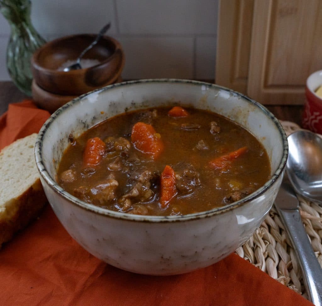 bowl of stew with a piece of bread on a cloth to the left side, spoon and knife to the right of the bowl 
