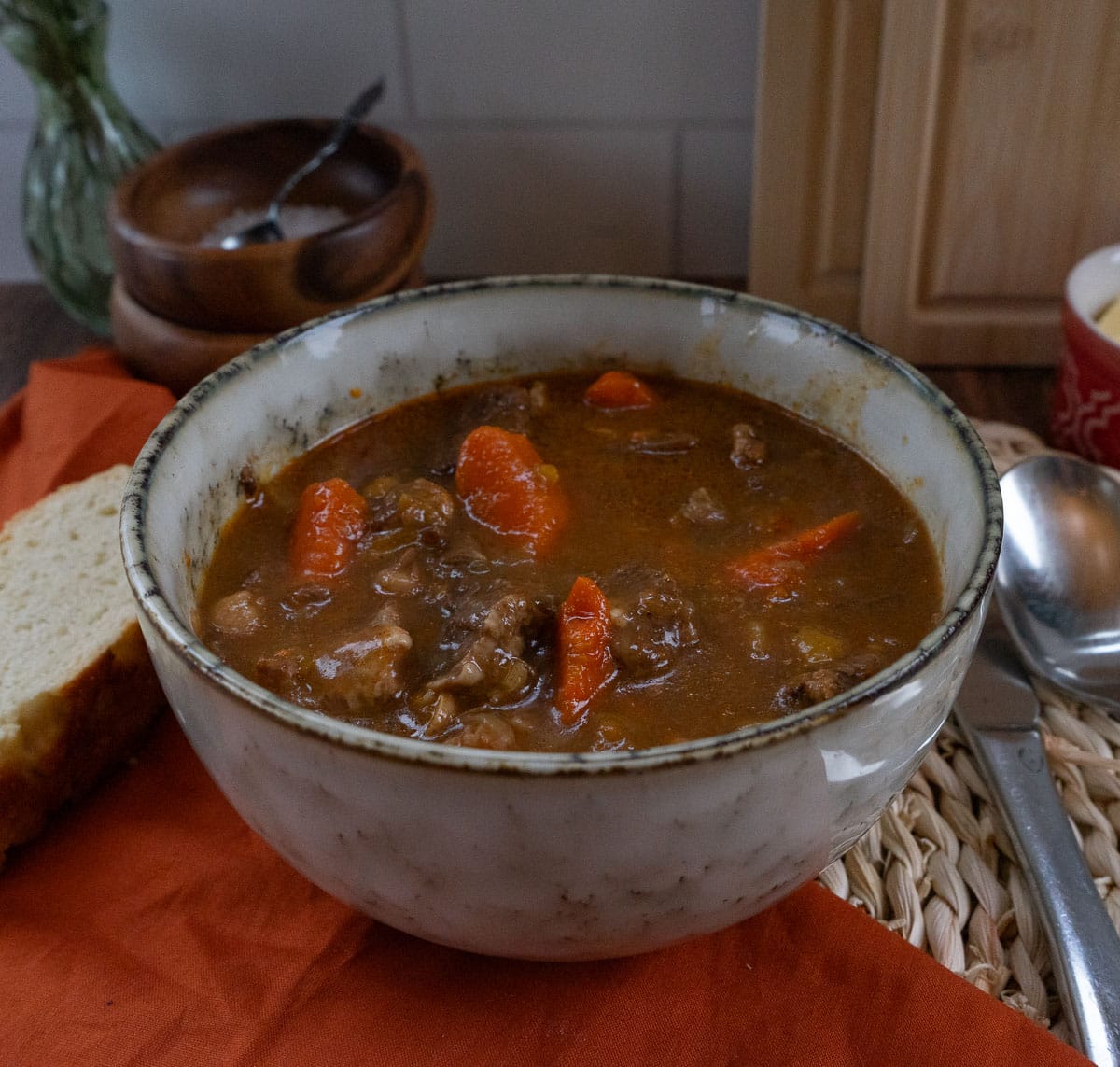 bowl of stew with a piece of bread on a cloth to the left side, spoon and knife to the right of the bowl
