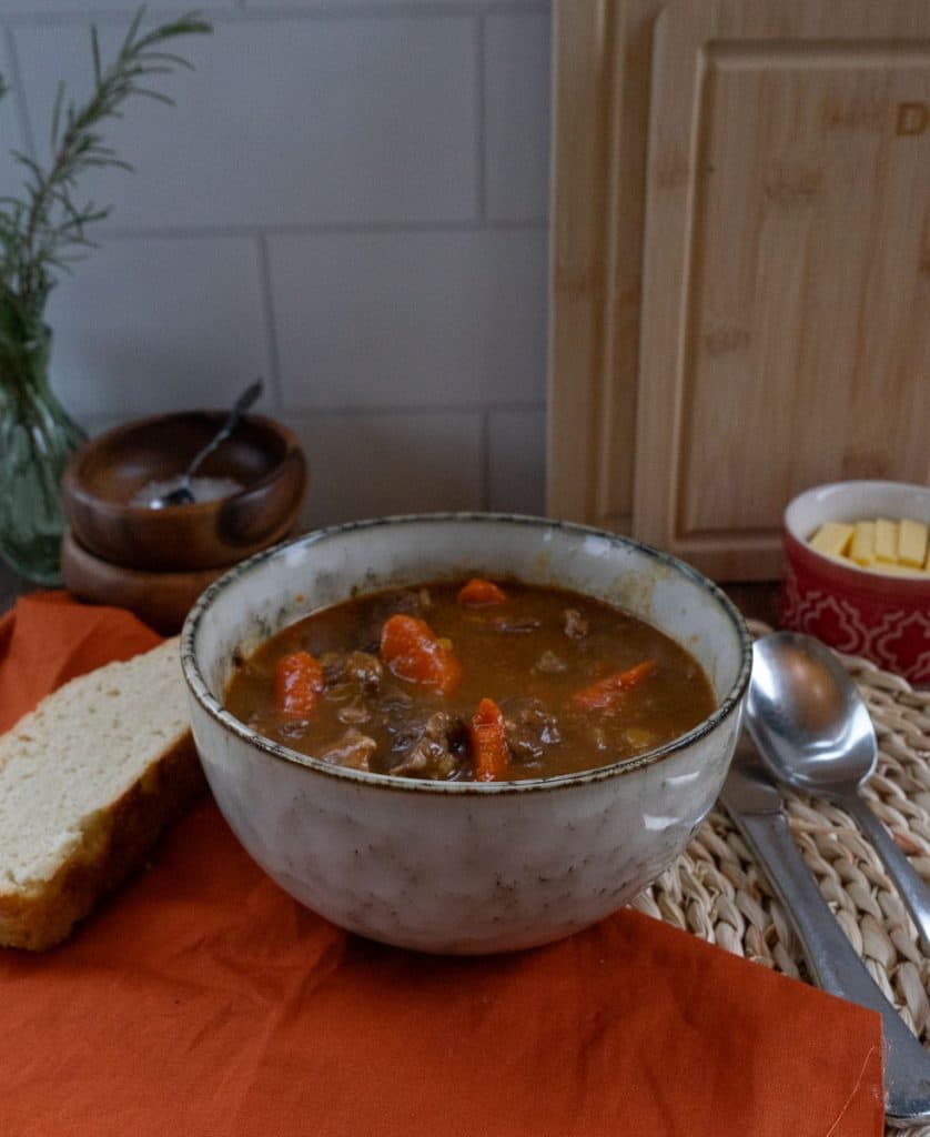 bowl of stew with a piece of bread on a cloth to the left side, spoon and knife to the right of the bowl