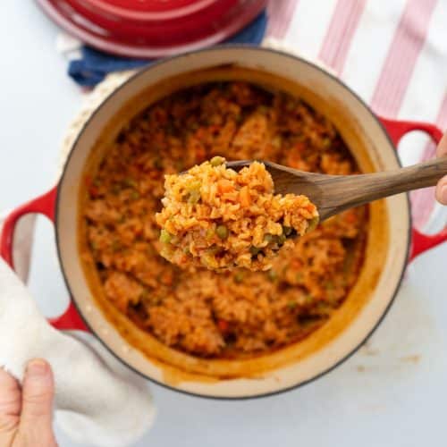 a pot of spanish rice with a hand scooping a spoonful out with a wooden spoon dutch oven is sitting on a red and white striped towel with the lid to the pot in the top half of the photo