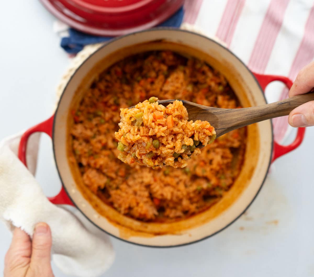 a pot of spanish rice with a hand scooping a spoonful out with a wooden spoon dutch oven is sitting on a red and white striped towel with the lid to the pot in the top half of the photo