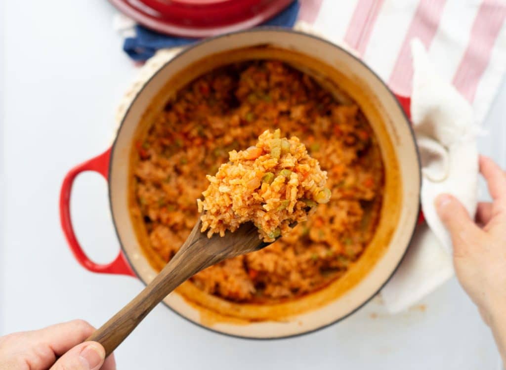 a pot of spanish rice with a hand scooping a spoonful out with a wooden spoon dutch oven is sitting on a red and white striped towel with the lid to the pot in the top half of the photo