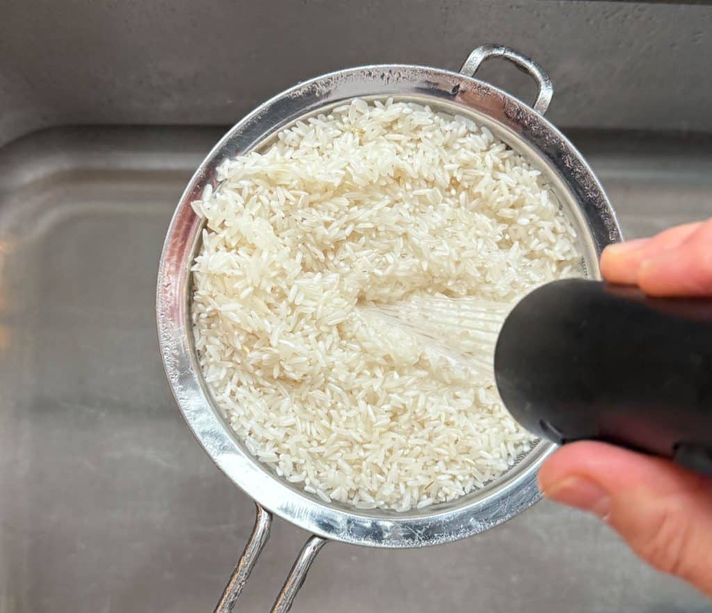 white long grain rice in a fine mesh sieve over a sink being rinsed