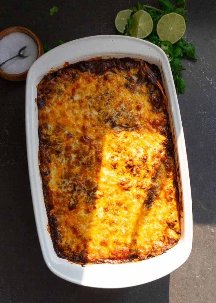 whole pan of easy layered beef and bean enchiladas on counter top with lime wedges and a wood bowl of salt in the top of the picture