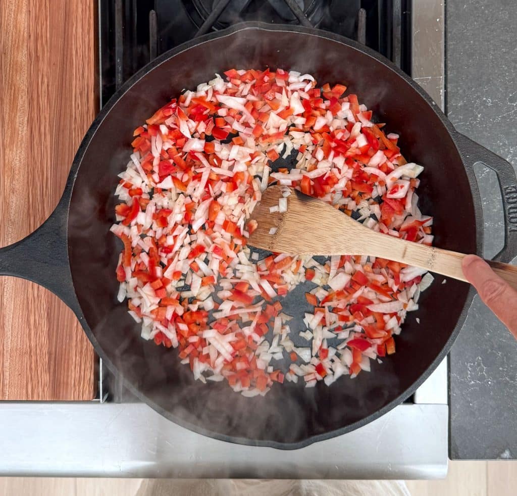 sauteing red bell peppers and onions in a cast iron skillet on the stove top with a wooden spoon in the pan