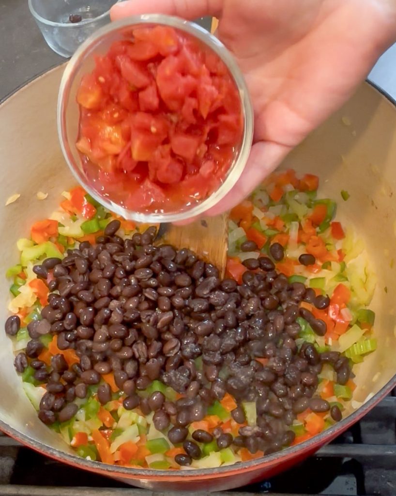 adding tomatoes to the other soup ingredients in a large pot on the stove top
