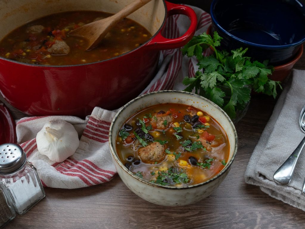 Bowl of mexican turkey meatball soup with a small bowl of cilantro behind it and the red pot of soup to the left