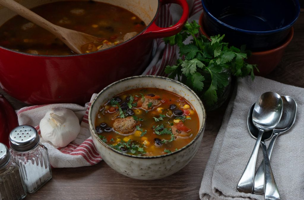photo of a bowl of mexican turkey meatball soup with a soup and napkin to the right of the bowl, cilantro and large pot of soup in the back fround salt and pepper shakers to the left of the bowl