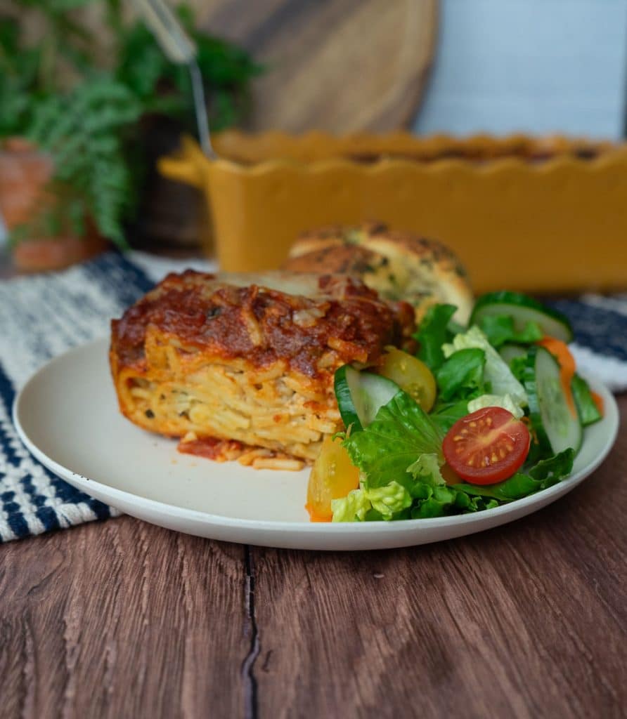 plate with a slice of spaghetti pie and side of salad and a garlic knot. the dish of spaghetti pie is in the background countertop is wood backsplash is white tile with a wood cutting board and a plant in the background
