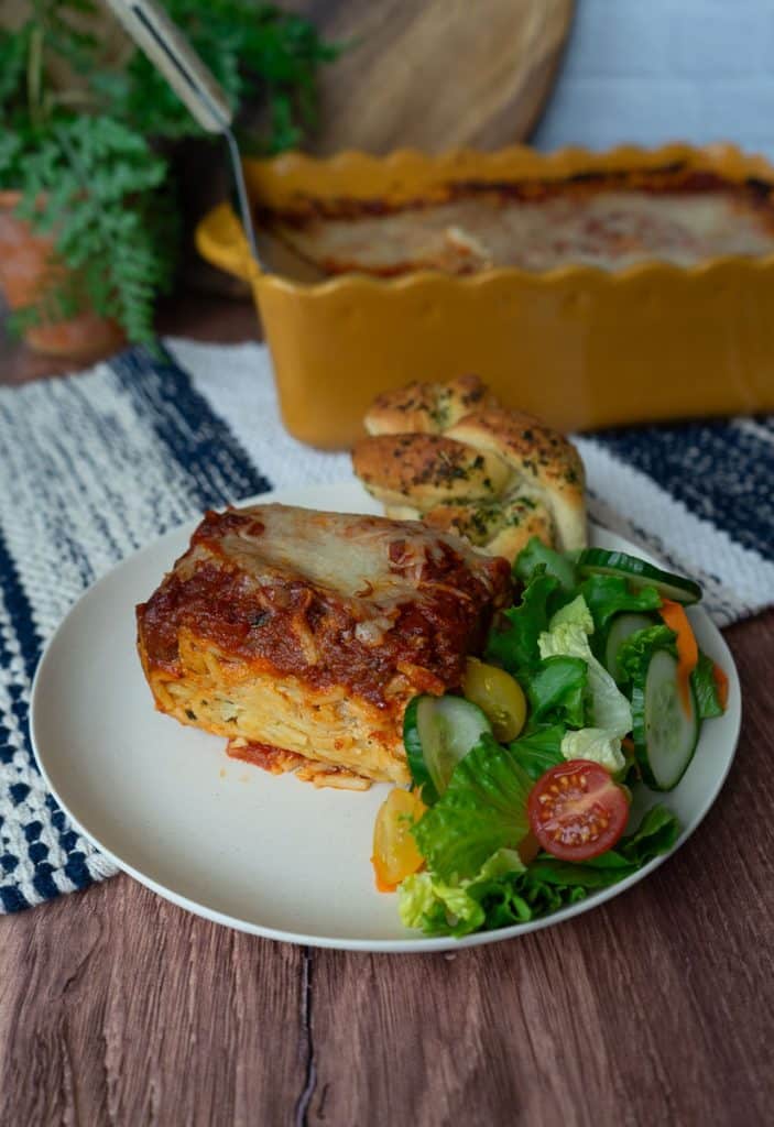 plate with a slice of spaghetti pie and side of salad and a garlic knot. the dish of spaghetti pie is in the background countertop is wood backsplash is white tile with a wood cutting board and a plant in the background