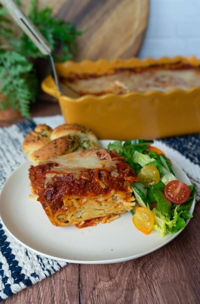 plate with a slice of spaghetti pie and side of salad and a garlic knot. the dish of spaghetti pie is in the background countertop is wood backsplash is white tile with a wood cutting board and a plant in the background