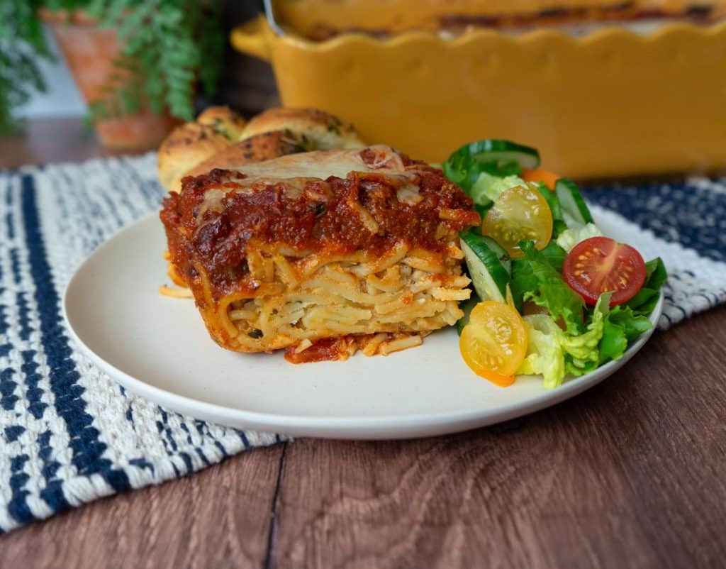 slice of spaghetti pie on a tan plate setting on a wood table with a blue and tan striped place mat dish of spaghetti pie is in the background