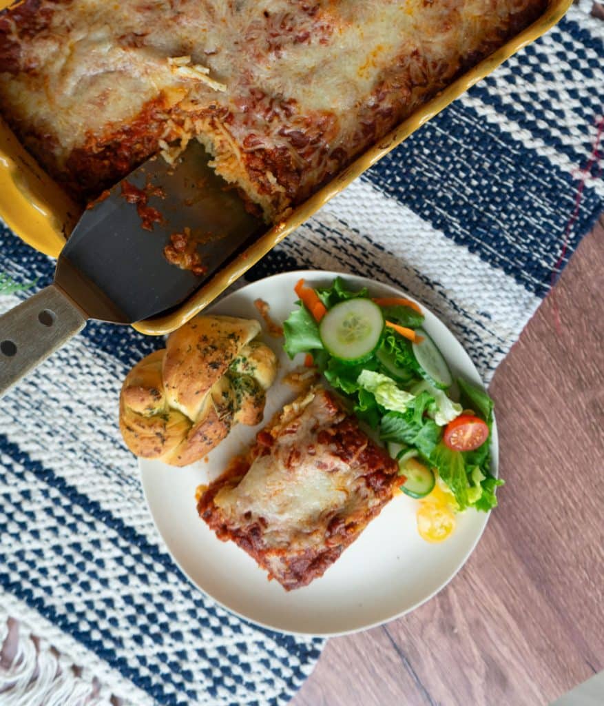top view of a plate with a slice of spaghetti pie and a salad with a garlic knot next to the full pan of pie. on a wood table with a red, white, and tan place matt